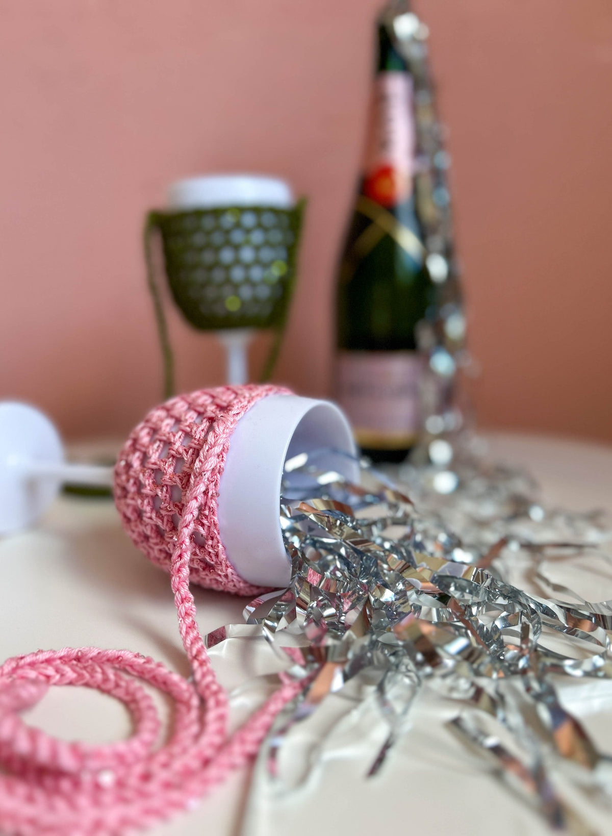Party Accessoires: rosa Weinglashalter mit Pailletten liegend im Lametta, dahinter Olivgrün und Sektflasche als Deko