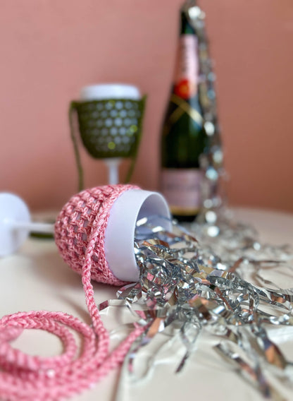 Party Accessoires: rosa Weinglashalter mit Pailletten liegend im Lametta, dahinter Olivgrün und Sektflasche als Deko