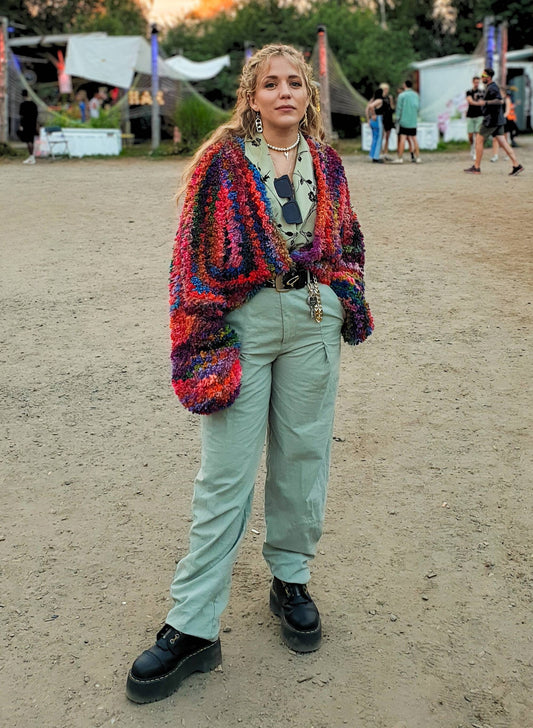 Frau stehr vor Festivalszene im Sonnenuntergang in buntem Merinocardigan in Schlaufenoptik